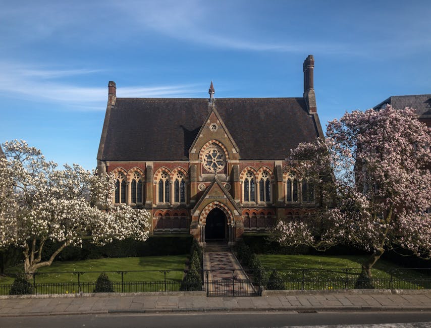 A large historic church building constructed with red and beige brickwork, featuring tall, narrow arched windows and a prominent circular stained glass window above the main entrance door. The church is surrounded by blooming trees with pink and white flowers, situated in a well-maintained green lawn enclosed by a low black metal fence. A paved walkway leads directly from the sidewalk to the church entrance, and the building is set against a clear blue sky with a few wispy clouds. The scene captures the exterior of the church during daytime, indicating a peaceful and picturesque environment that might be relevant to local community or relocation services such as those offered by Harrow Movers in Wealdstone near Harrow.