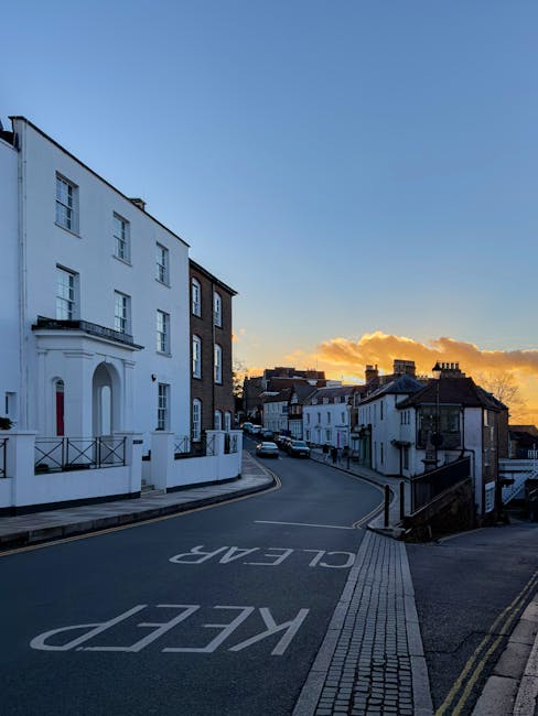 A quiet residential street in Wealdstone near Harrow at sunset, showcasing a gentle slope with a variety of white and dark brick terraced houses lining the road. The pavement features tactile paving strips near the curb, while a large white 'KEEP CLEAR' marking is painted on the asphalt, indicating a no-parking zone. In the foreground, a section of the pavement is visible, with some small metallic bollards and a black metal railing along the stairs descending to a lower street level. The sky is partly cloudy with orange and yellow hues from the setting sun, casting a warm glow over the scene. The street is relatively empty, with a few parked cars further down, and no visible activity related to house removals or furniture transport. The overall scene offers an unobstructed view of the typical street environment where home relocation services by Harrow Movers could be performed, with an emphasis on the residential setting and the infrastructural elements involved in moving logistics.