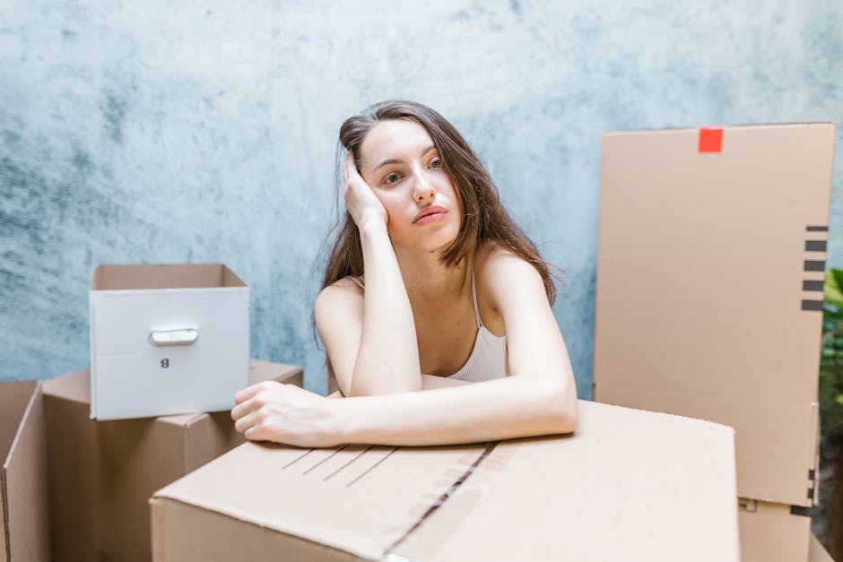 A young woman with long brown hair, wearing a white tank top, rests her head on her hand with a bored or exhausted expression, surrounded by several large cardboard boxes, some sealed with packing tape and others open, in an indoor space with a plain light blue wall. The boxes, made of brown corrugated cardboard, vary in size and are stacked or placed on a sliding surface, possibly during a home relocation or packing process. The lighting is natural and evenly distributed, highlighting the cluttered environment common during household moving or furniture transport activities. This scene reflects the logistical challenges and stress often associated with packing and moving, as depicted by the woman's fatigued demeanor amidst the boxes, with Harrow Movers providing professional removals and moving services connected to this process.