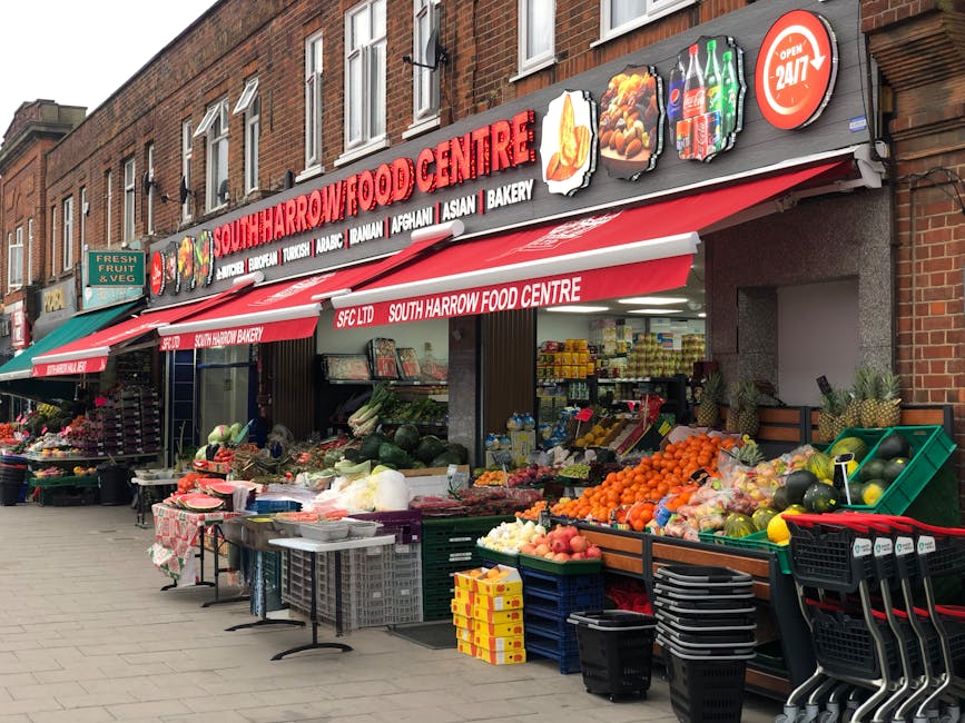 A street-level view of South Harrow Food Centre, a grocery store with a red and white signage displaying the store name and various food categories, including burger, Turkish, Afghan, Asian, and bakery. The store has a red awning with the text 'South Harrow Food Centre' repeating along its length. Outside the shop, several green and black plastic crates and baskets are filled with an assortment of fresh produce such as oranges, watermelons, apples, and vegetables, arranged on wooden display tables and stacked on the pavement. Some produce is wrapped in transparent plastic, and others are loose, with a scale and a small cash register also visible. To the left, additional fruits and vegetables are arranged on a tiered stand, with a small green sign indicating fresh fruit and vegetables. The environment appears to be late morning or early afternoon, with natural daylight illuminating the scene, and the setting is part of the outdoor area of a building that could be used for home relocation or moving logistics, supporting services like packing or transport with Harrow Movers.