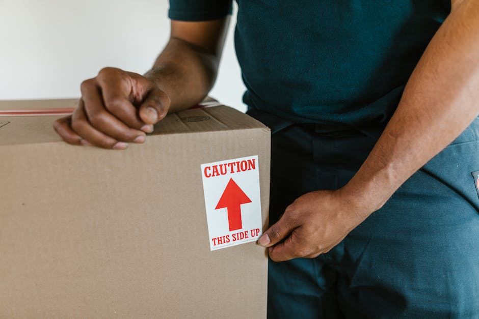 A young woman with long brown hair, wearing a white tank top, rests her head on her hand with a bored or exhausted expression, surrounded by several large cardboard boxes, some sealed with packing tape and others open, in an indoor space with a plain light blue wall. The boxes, made of brown corrugated cardboard, vary in size and are stacked or placed on a sliding surface, possibly during a home relocation or packing process. The lighting is natural and evenly distributed, highlighting the cluttered environment common during household moving or furniture transport activities. This scene reflects the logistical challenges and stress often associated with packing and moving, as depicted by the woman's fatigued demeanor amidst the boxes, with Harrow Movers providing professional removals and moving services connected to this process.