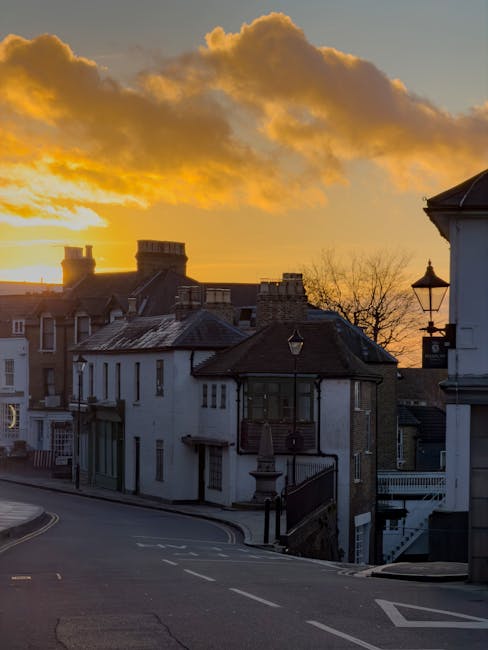 A street scene during sunset in Harrow on the Hill featuring a row of residential buildings with varied architecture, including a white building with a small balcony and black railings, and another with a bay window. The sky is illuminated with orange and yellow hues from the setting sun, with large, partly cloudy clouds above. In the foreground, a paved road curves gently, with a pedestrian crossing and road markings visible. To the right, a lamppost stands beside a building with a dark roof, suggesting an urban area in the process of home relocation or moving services. Harrow Movers, a professional removals company, may facilitate the transportation of furniture and household goods from such properties, supporting a smooth moving process within the Harrow district.