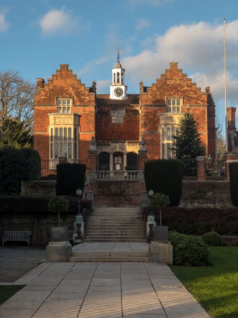 The image shows the front facade of a historic brick building with a central clock tower and a decorative gable roof, part of Harrow on the Hill in London. The building features large, ornate windows with stone detailing, a staircase leading up to the entrance, and a well-maintained garden area with trimmed bushes, a bench, and paved pathways. The scene is illuminated by natural daylight, with a clear sky and some clouds overhead. This setting reflects a typical location for house removals and apartment relocations, with the building exterior providing context for the types of properties serviced by Harrow Movers during furniture transport, packing, and home relocation processes.