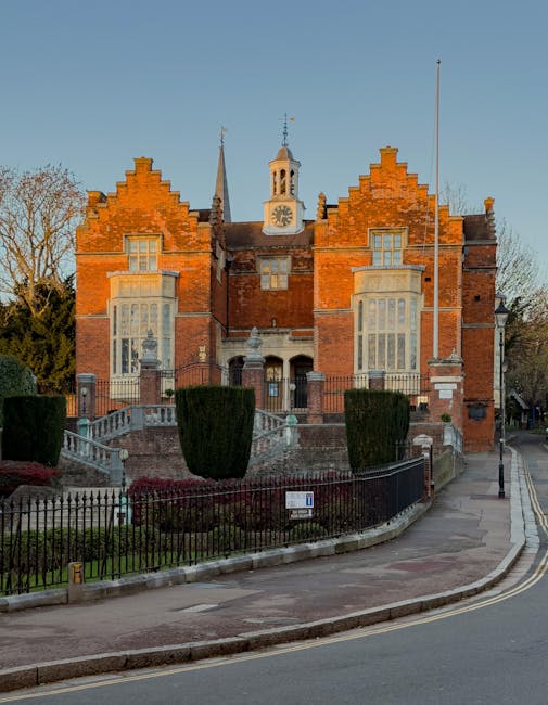 A historic red-brick building with decorated gable ends, tall narrow windows, and a central clock tower topped with a weather vane, situated on a paved street. In front of the building, there is a sma