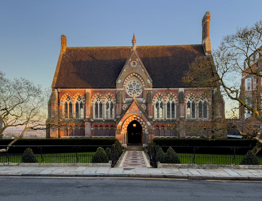 A historic red-brick building with decorated gable ends, tall narrow windows, and a central clock tower topped with a weather vane, situated on a paved street. In front of the building, there is a sma