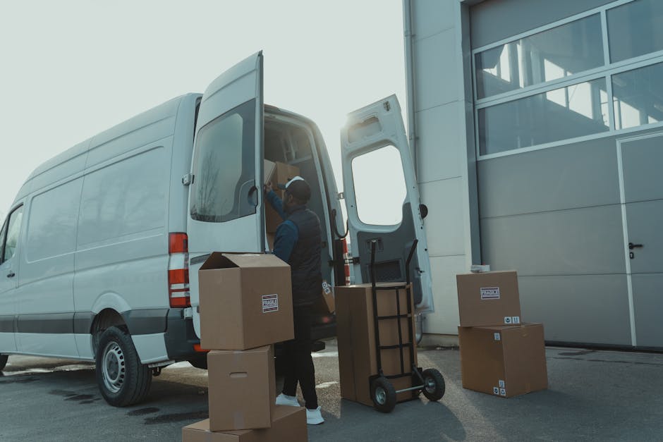 A warehouse or loading dock area during daylight with a white delivery van positioned outdoors near a modern building with large windows. The side door of the van is open, revealing a person wearing a cap, vest, and casual clothing, loading or arranging cardboard boxes and packing materials inside. Several sealed cardboard boxes, some with shipping labels, are arranged on the ground and stacked nearby. A hand truck or dolly with additional boxes is positioned adjacent to the van, ready for transporting items. The environment is well-lit by natural sunlight, and the scene captures a typical home relocation or furniture transport process characteristic of professional removals services, such as those offered by Harrow Movers.