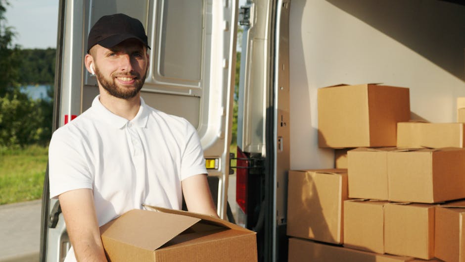A warehouse or loading dock area during daylight with a white delivery van positioned outdoors near a modern building with large windows. The side door of the van is open, revealing a person wearing a cap, vest, and casual clothing, loading or arranging cardboard boxes and packing materials inside. Several sealed cardboard boxes, some with shipping labels, are arranged on the ground and stacked nearby. A hand truck or dolly with additional boxes is positioned adjacent to the van, ready for transporting items. The environment is well-lit by natural sunlight, and the scene captures a typical home relocation or furniture transport process characteristic of professional removals services, such as those offered by Harrow Movers.