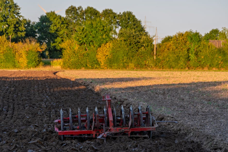A red agricultural tiller attachment is positioned on freshly tilled dark brown soil in an open field, near the edge of a paved area. The tiller has multiple metal blades and tines, and is connected t