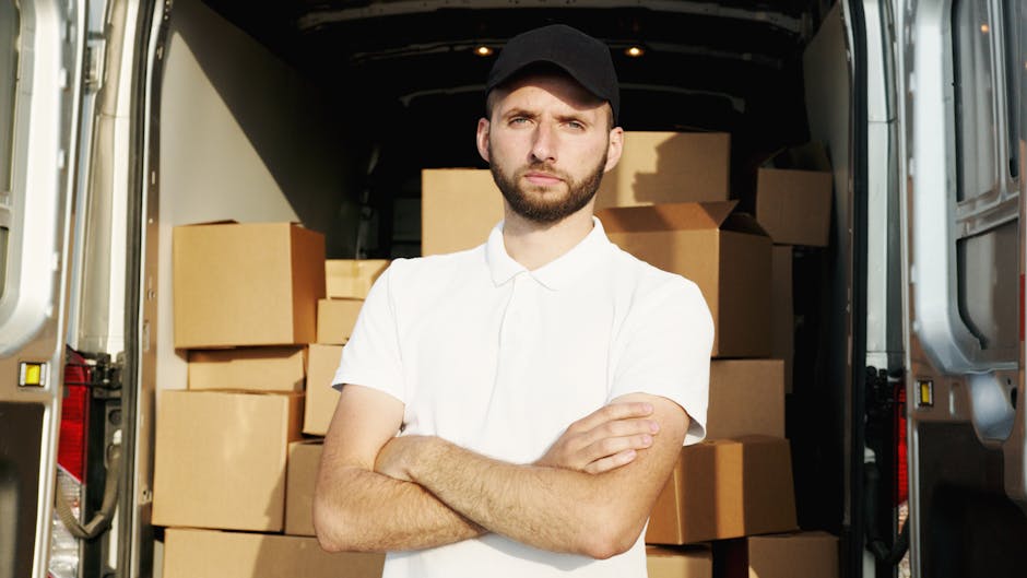 A man with a serious expression, wearing a white polo shirt and a black cap, stands inside the open rear doors of a white moving van filled with neatly stacked cardboard boxes of various sizes for a house relocation. The boxes are sealed with packing tape and are arranged on the vehicle's floor, some leaning against the walls, with a few smaller boxes placed on top of larger ones. The environment is well-lit, with natural light illuminating the interior of the van and the man's face. Visible move equipment includes a metal trolley partially seen on the left side. Outside, the van appears to be parked on a paved area, likely in a driveway or on the street. This scene represents the loading process of furniture transport and packing during home moving services, as undertaken by a professional removals company like Harrow Movers, involved in same day removals in Harrow.